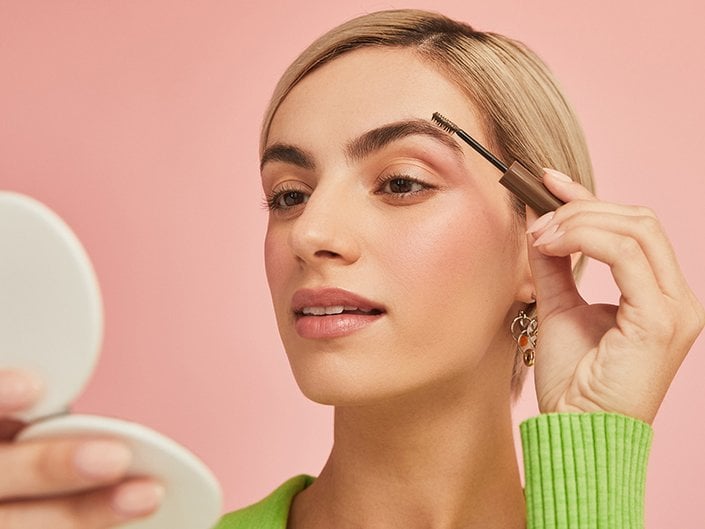 Person with a blonde pixie cut applying brow gel with a spoolie brush applicator to their eyebrow. 