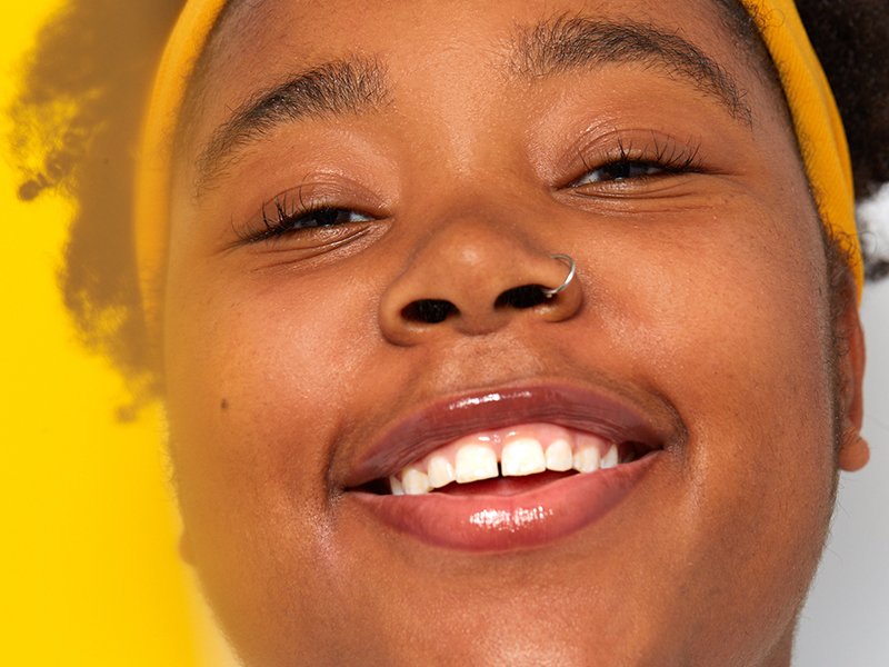 Close-up picture of a person with a silver nose ring smiling, against a bright yellow background