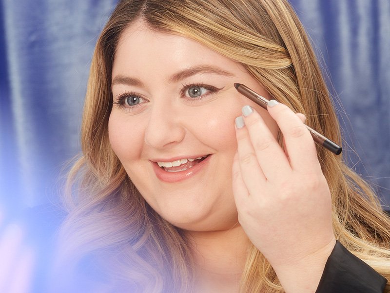 Picture of a blonde person with hooded eyes smiling and holding an eyeliner pencil to their eye