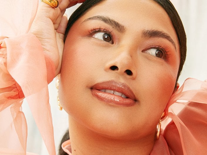 A person with their dark hair pulled back wears orange-toned blush and glossy lips