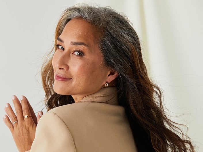 Person with long, wavy graying hair looking over their shoulder at the camera and smiling with their mouth closed