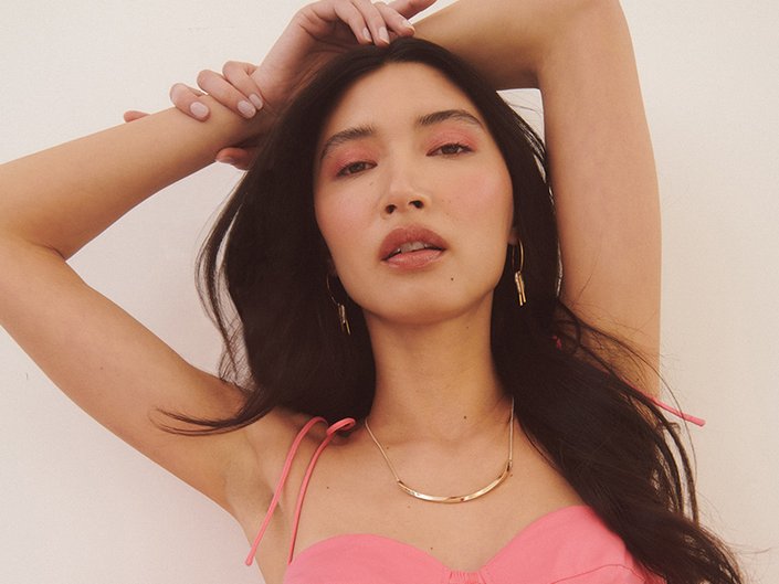 Model with long dark hair wearing pink eyeshadow, gold hoop earrings, a gold necklace and a pink top