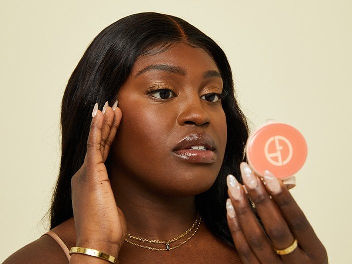 Photo of a person with long dark hair and long nails holding their fingers to their temple and looking in the mirror of a Giorgio Armani Beauty compact