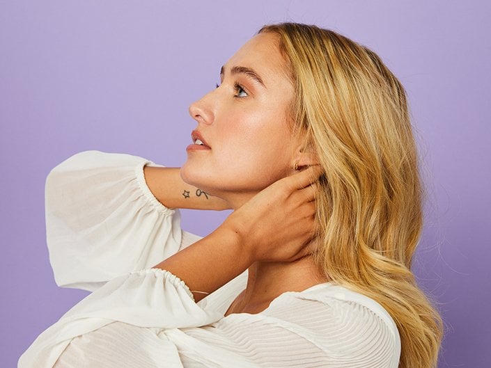 Photo of person with long blonde hair in profile holding their hands on their neck, standing against a purple background