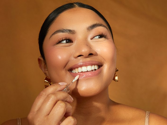 Person with their dark hair pulled back, smiling and applying lip gloss