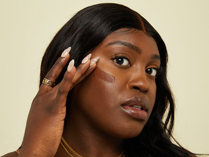 Photo of a Black model looking at the camera and swatching foundation on their cheek