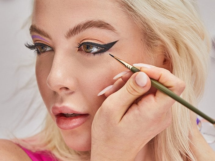 Person with light blonde hair and long nails using an eyeliner brush to apply a black winged eyeliner look