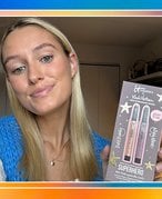Photo of editor with long blonde hair wearing light makeup and a blue knit cardigan, holding up the IT Cosmetics x Madi Nelson Superhero No-Tug Eyeshadow Sticks Kit. The photo is collaged on a rainbow tie-dye background.