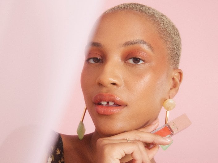 Picture of a model with bleached blonde buzzcut holding a lip product on a soft pink background