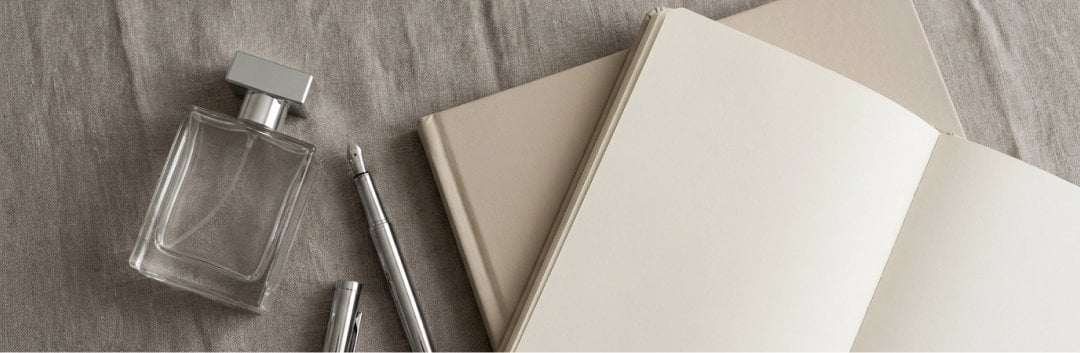 A clean, minimalist flat lay on a warm gray linen surface. A single modern perfume bottle sits next to a sleek silver fountain pen and an open minimalist planner, captured under soft, diffuse daylight with gentle, clean shadows.