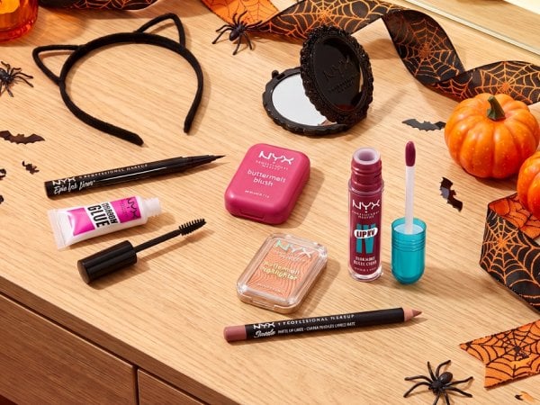 NYX Professional Makeup Epic Ink Liner, Brow Glue Instant Brow Styler, Buttermelt Blush, Buttermelt Highlighter, Lip IV Hydrating Gloss Stain, and Suede Matte Lip Liner arranged on a Halloween vanity with a compact mirror, cat-ear headband, spiderweb ribbon, and black-and-orange props. No people or faces.