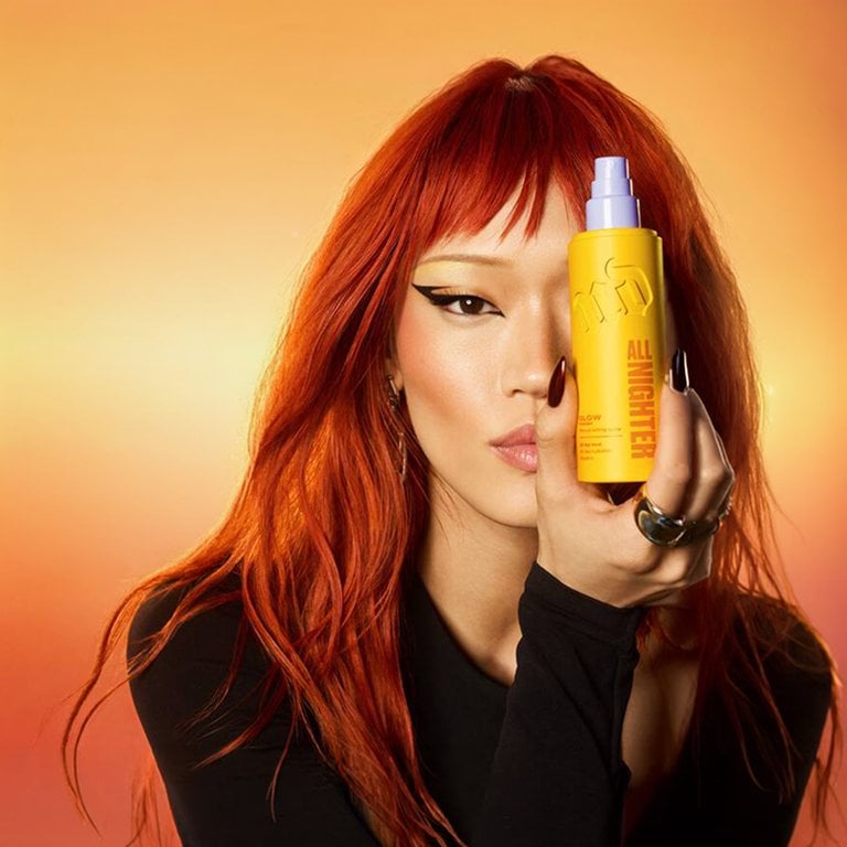 Woman with red hair holding a bottle of Urban Decay All Nighter Glow Waterproof Setting Spray