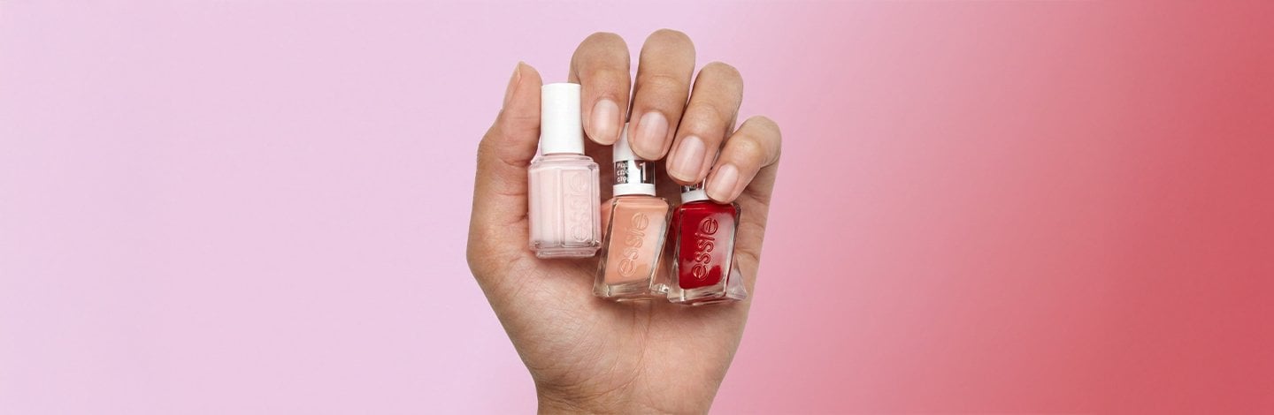 A hand gripping three bottles of Essie nail polish in the shades Ballet Slippers, Of Corset, and Bubbles Only on a pink background