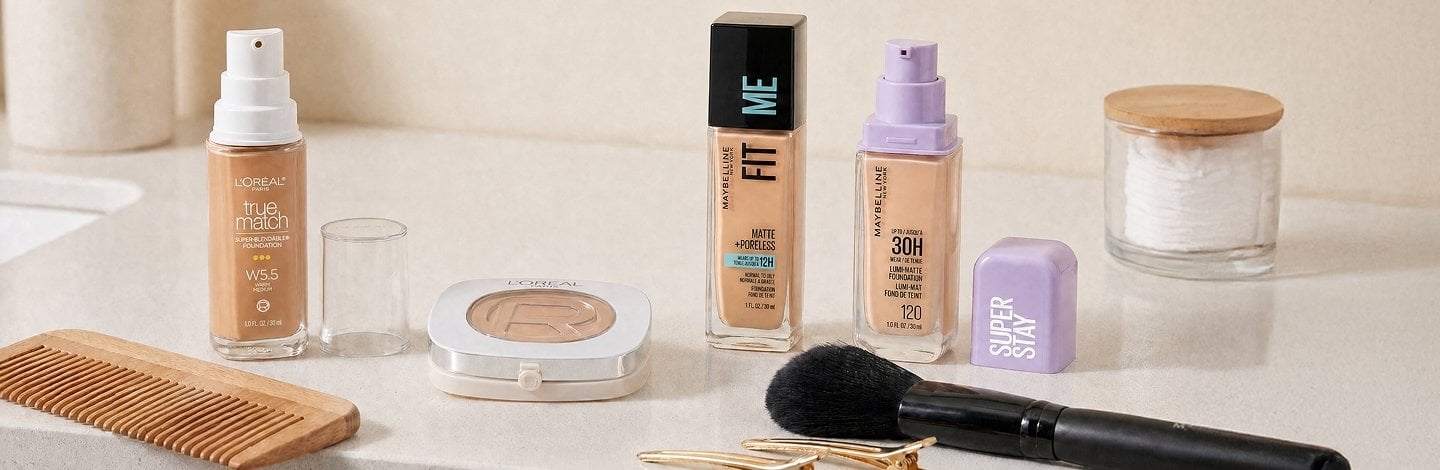 A bathroom counter displaying some of the foundation products mentioned in this article: L’Oréal Paris True Match Super-Blendable Foundation, L’Oréal Paris True Match Hyaluron Tinted Balm, Maybelline Fit Me Matte + Poreless Foundation, Maybelline Super Stay Lumi-Matte Foundation, comb/brush, hair clips, makeup brush, etc.