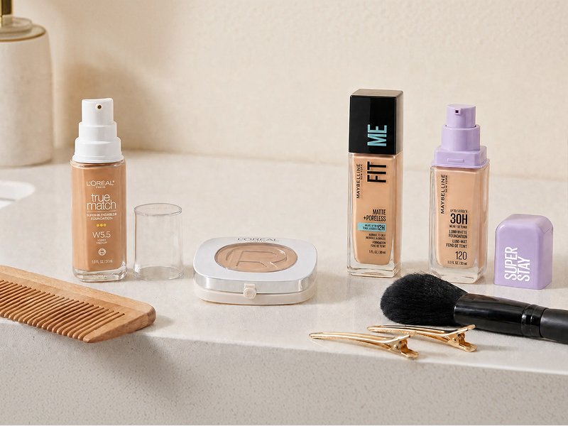 A bathroom counter displaying some of the foundation products mentioned in this article: L’Oréal Paris True Match Super-Blendable Foundation, L’Oréal Paris True Match Hyaluron Tinted Balm, Maybelline Fit Me Matte + Poreless Foundation, Maybelline Super Stay Lumi-Matte Foundation, comb/brush, hair clips, makeup brush, etc.