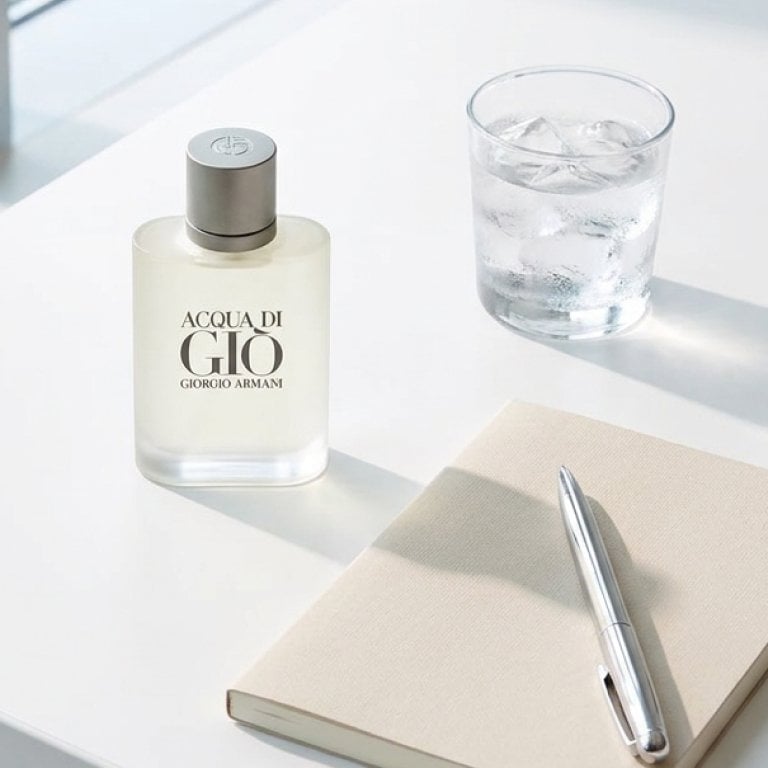 A bright, airy flat lay on a clean white desk, featuring a sleek bottle of Acqua di Giò Eau de Toilette next to a neatly arranged notepad, a silver pen, and a small glass of ice water, all illuminated by soft, natural daylight streaming through a clean office window. 