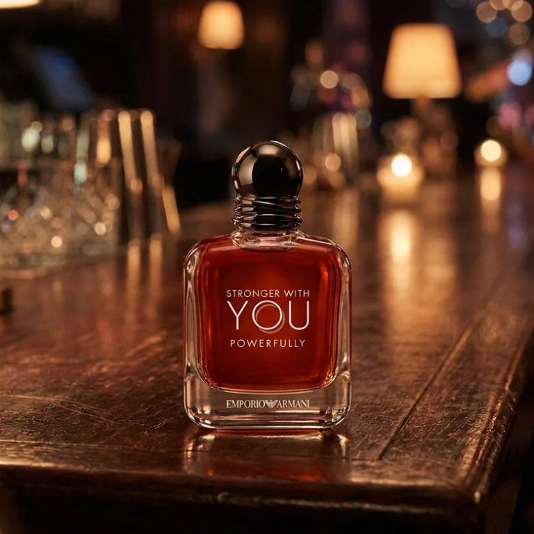 A moody, atmospheric bar scene captured in warm, dim lighting, featuring a sleek bottle of Stronger With You Powerfully Eau de Parfum resting on a dark polished wooden table next to a crystal glass of amber-toned spirits, with a soft-focus background of ambient evening lights and lively social activity.