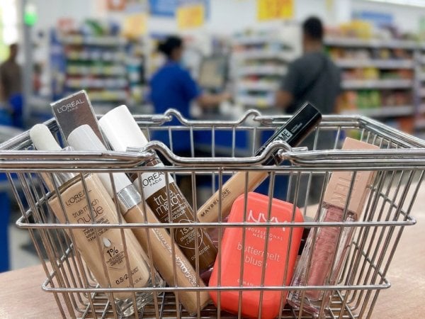 a basket at the counter with the following items: L'Oréal Paris True Match Nude Hyaluronic Tinted Serum, L'Oréal Paris True Match Radiant Serum Concealer, Maybelline New York Fit Me Concealer, NYX Professional Makeup Buttermelt Blush, Maybelline New York Lifter Gloss, Focus on the products. The background hypermart scene is blurred out.