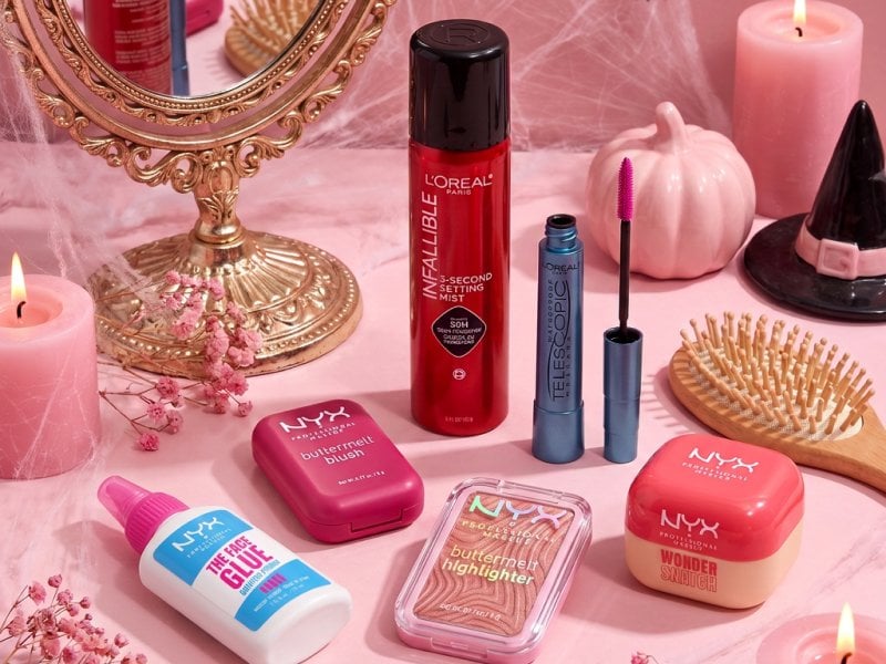 Long-wear Halloween makeup products, including NYX Professional Makeup Pore Filler Primer, Wonder Snatch Loose Setting Powder, Buttermelt Blush, Buttermelt Highlighter, L’Oréal Paris Telescopic Original Waterproof Mascara, and L’Oréal Paris Infallible 3-Second Makeup Setting Spray Mist arranged on a Halloween vanity. No people or faces. 